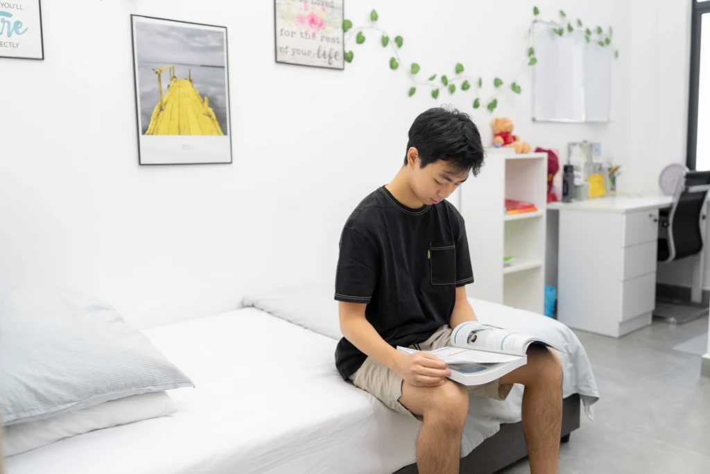 A boarder reads a book while sitting on the bed in their personalised room.