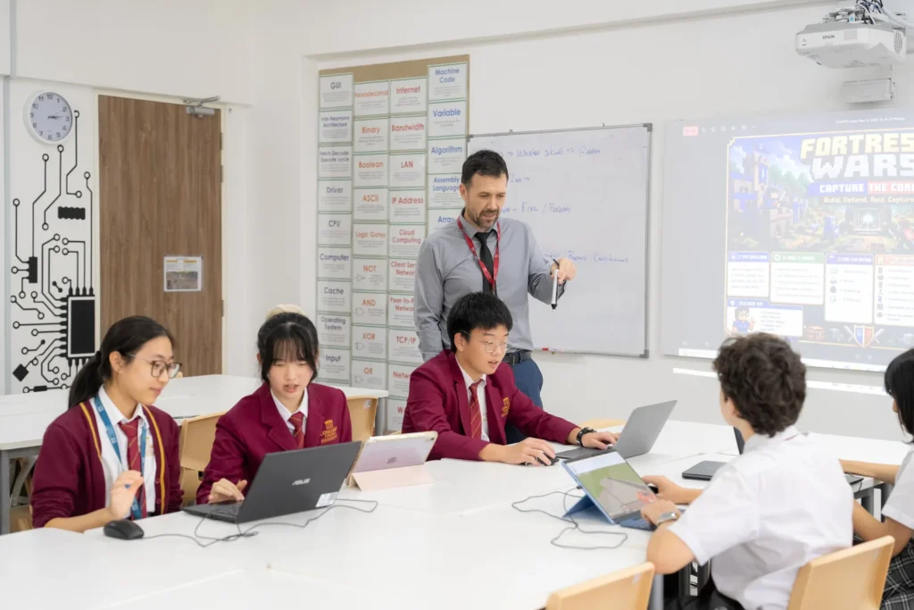 Secondary students working at laptops during a computing lesson while their teacher leads from the front.