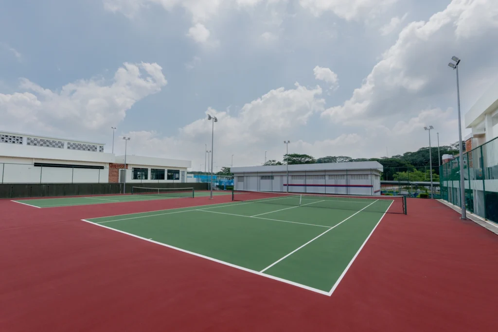 An outdoor tennis court with green playing surface bordered by red, surrounded by white buildings under a partly cloudy sky.