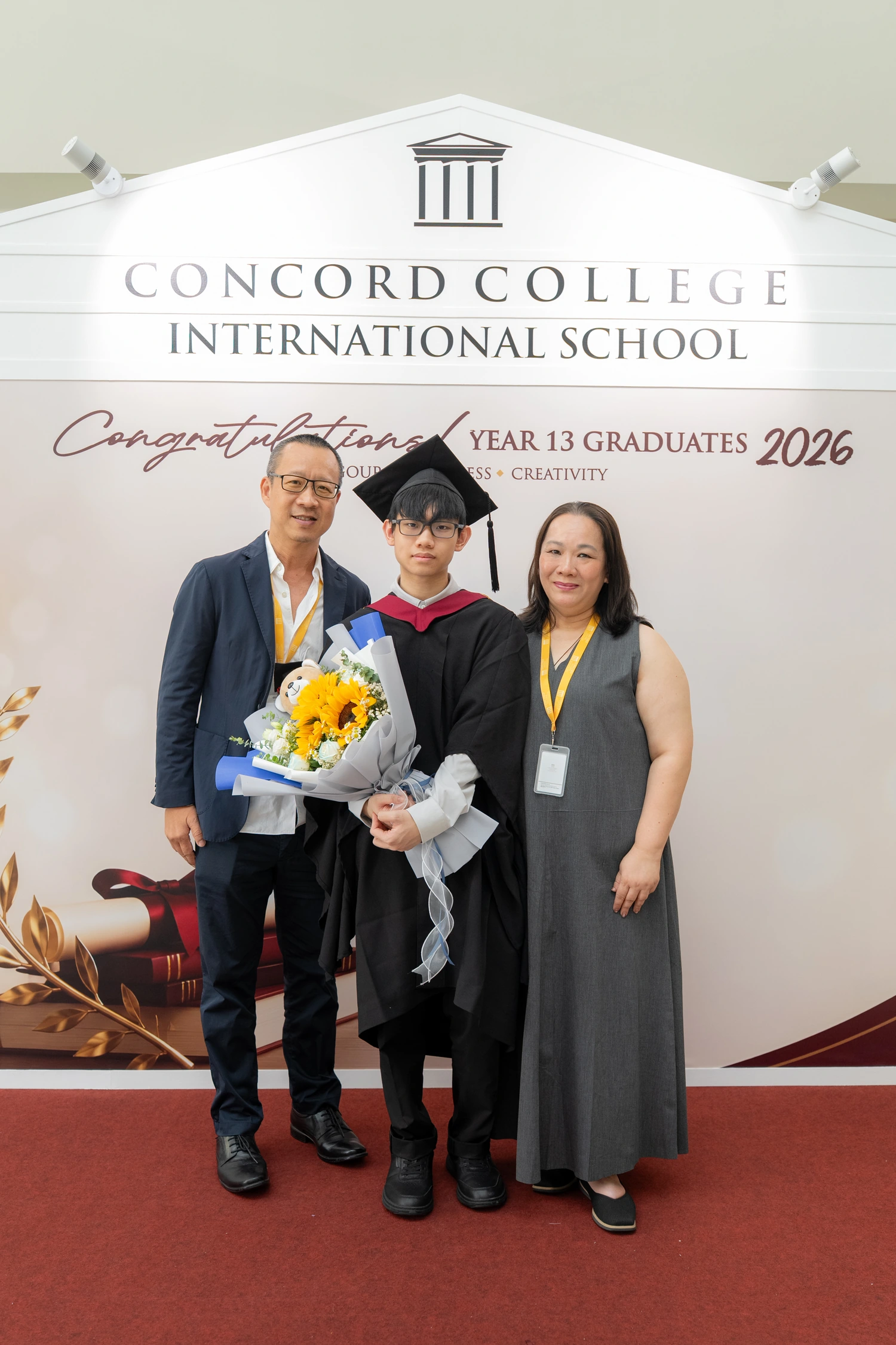 A graduate celebrates with family and a bouquet at the graduation backdrop.