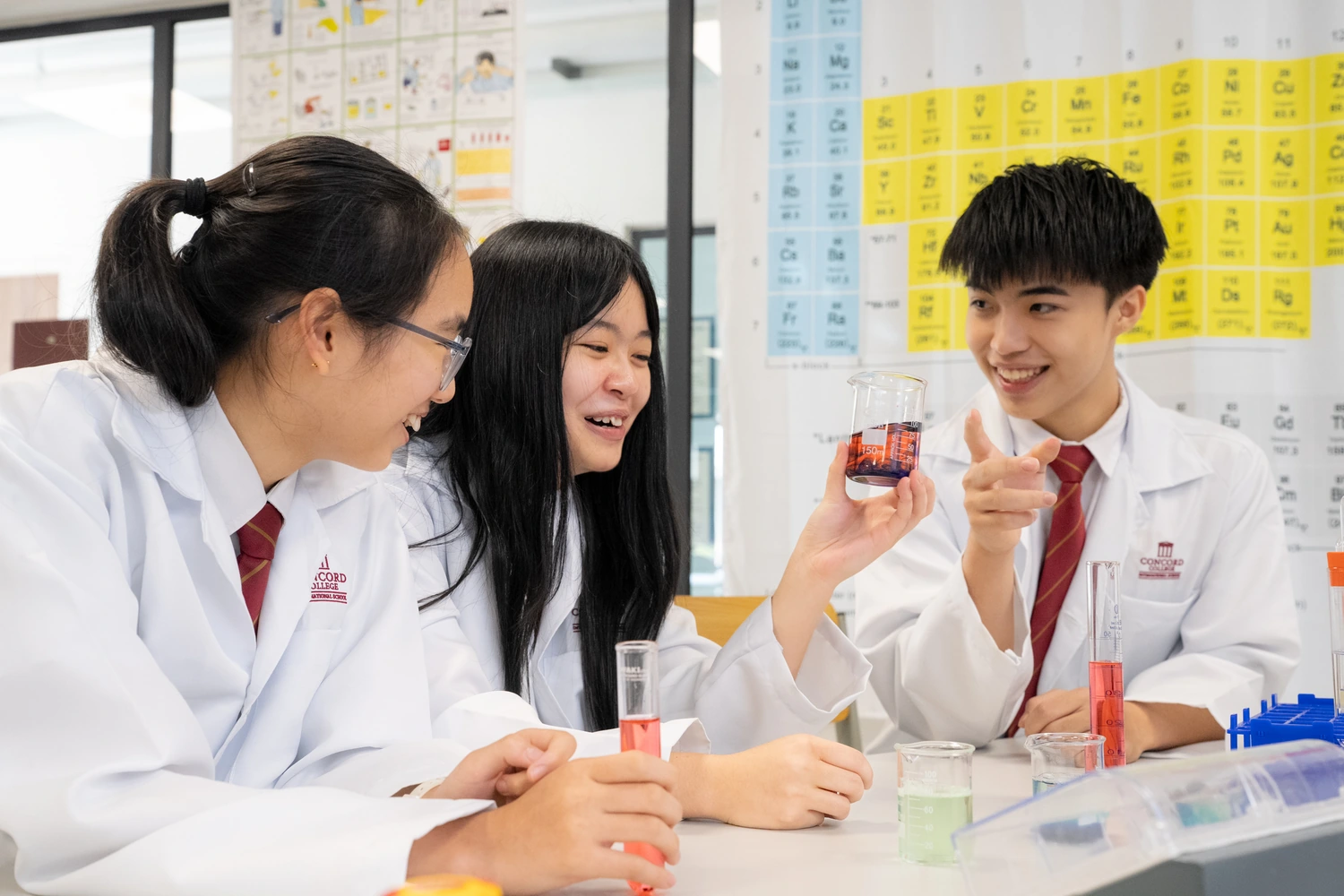 Three students in lab coats hold up test tubes of coloured solution in the chemistry laboratory.