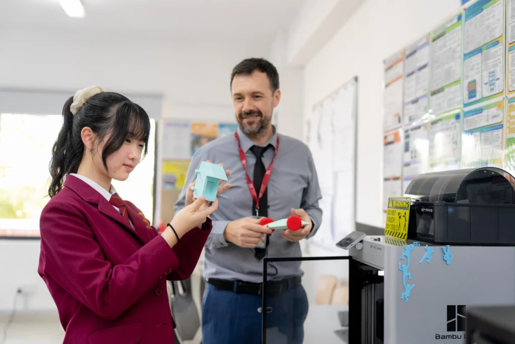 A student shows a 3D-printed model to a teacher beside the 3D printer.