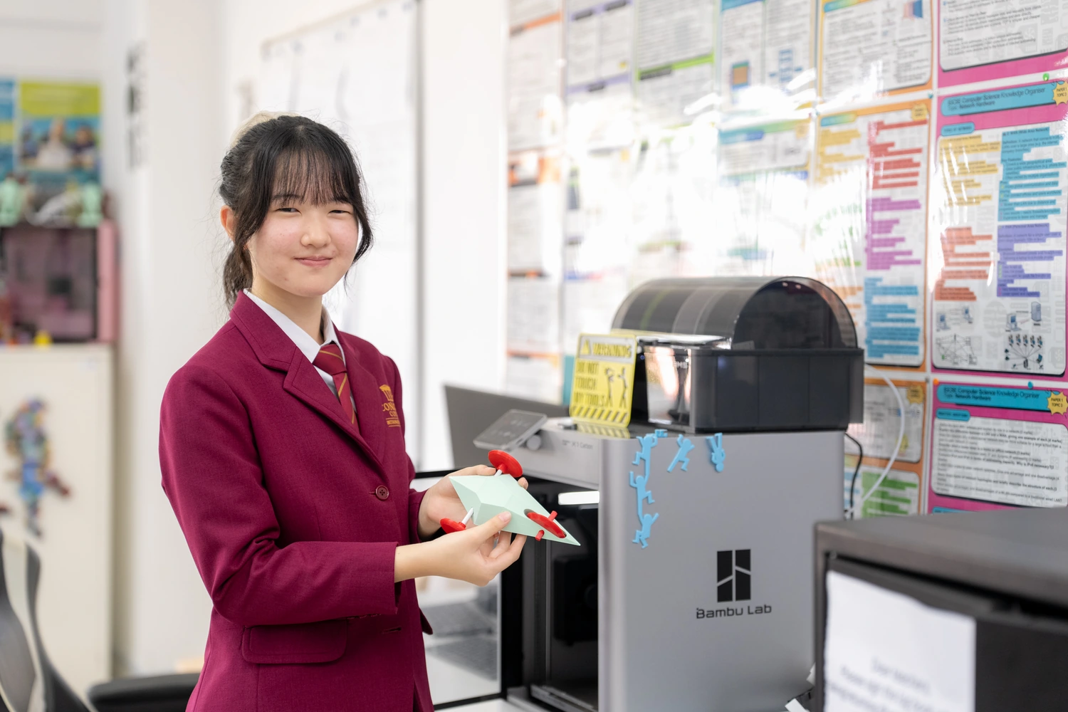 A student holds up a 3D-printed model beside the 3D printer in the computing room.