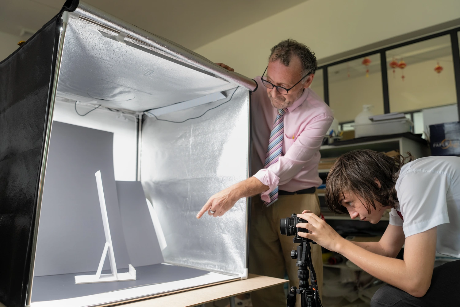 A teacher and a student photograph a model inside a lighting tent in the design and technology room.