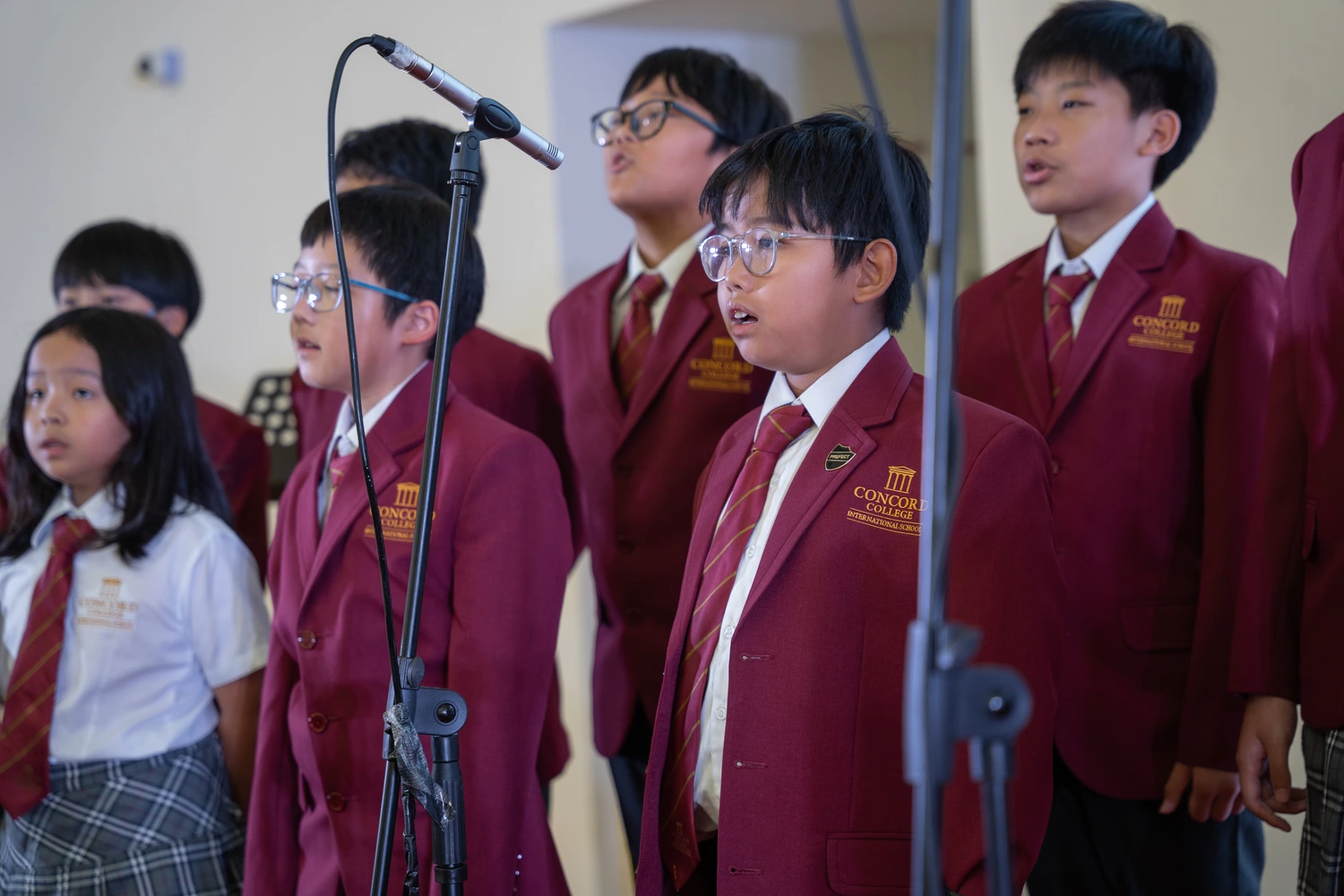 Students in school blazers singing together around a microphone stand.