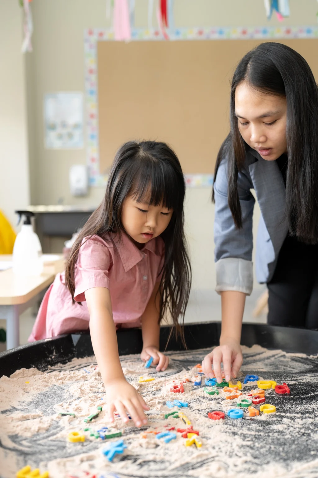 A young girl and an adult work with colourful letters on a sand tray in the Early Years classroom.