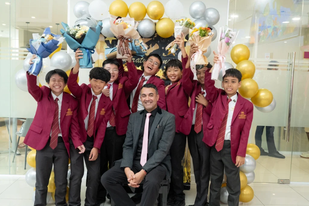 Year 6 graduates in maroon blazers cheer with bouquets around their teacher at the graduation celebration.